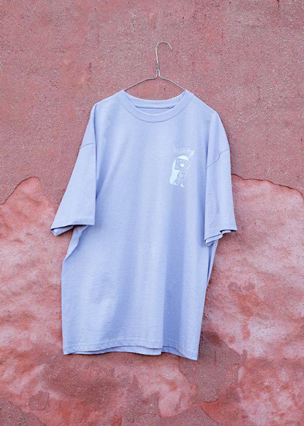 A light purple T-shirt hangs on a wire hanger against a textured, reddish-pink wall. The T-shirt features a small graphic design on the chest.