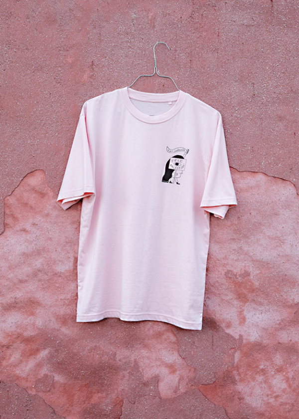 A light pink T-shirt hanging on a wire hanger against a textured pink wall. The shirt has a small black and white graphic on the front featuring a cartoon-like character.