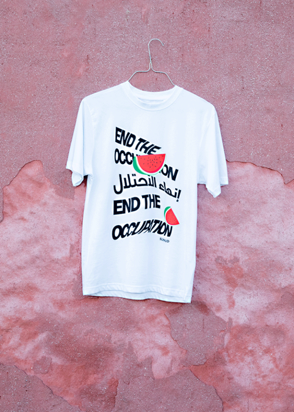 A white T-shirt hangs against a textured pink wall. The shirt features the text "End the Occupation" in both English and Arabic, accented with illustrations of watermelon slices.