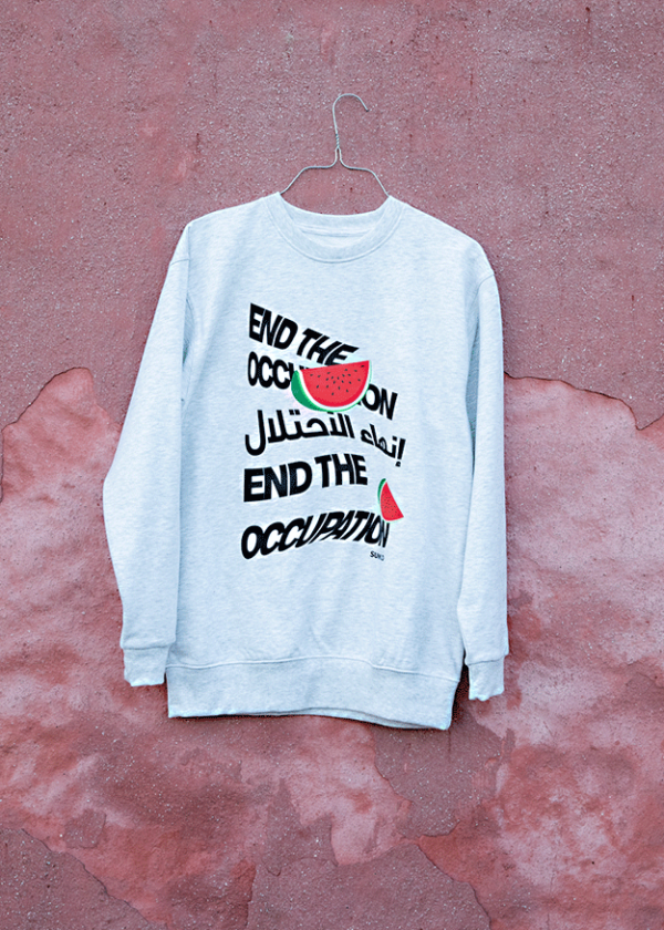 A gray sweatshirt hangs against a distressed pink wall. The shirt features bold, stylized text reading "END THE OCCUPATION" in English and Arabic, with watermelon illustrations integrated into the design.