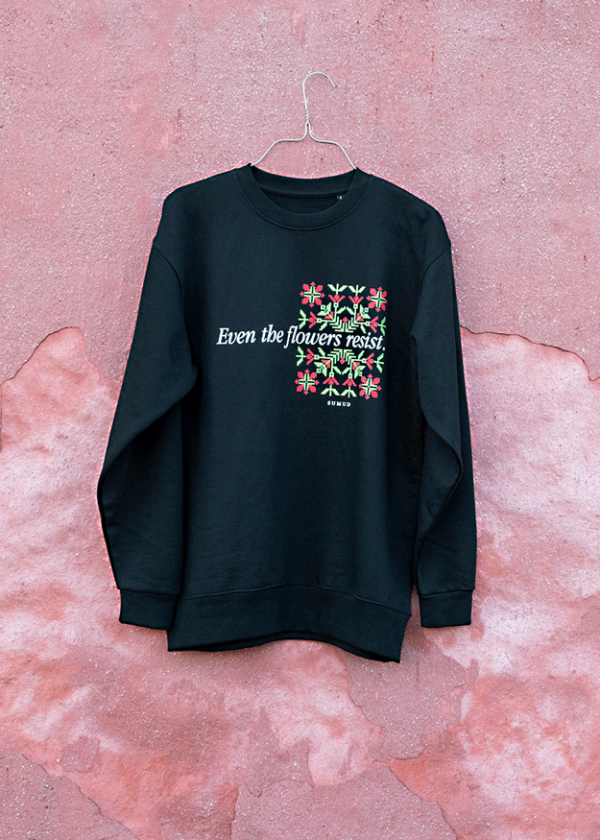 A black sweatshirt hangs on a hanger against a textured pink wall. It features a floral design and the text "Even the flowers resist" in white.