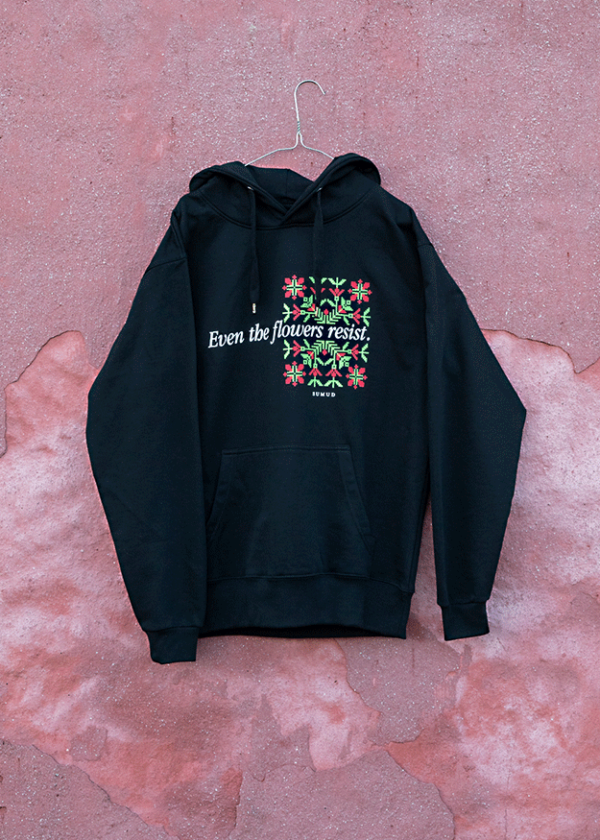 A black hoodie hangs on a hook against a pink textured wall. The hoodie features colorful embroidered flowers and the text "Even the flowers resist" across the chest.