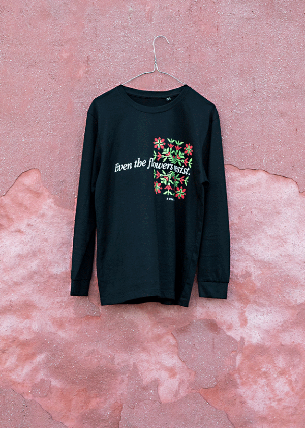 A black long-sleeve shirt is hanging on a wire hanger against a textured pink wall. The shirt features floral designs and the text "Even the flowers resist" in white lettering.