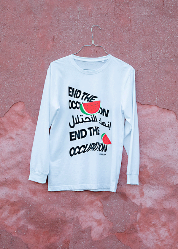 A white long-sleeve shirt hanging against a textured pink wall. The shirt features text in English and Arabic stating "End the Occupation" alongside a red and green watermelon graphic.