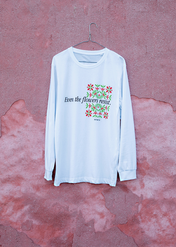 A white long-sleeve shirt hangs on a pink textured wall. The shirt features colorful floral designs and the text "Even the flowers resist.