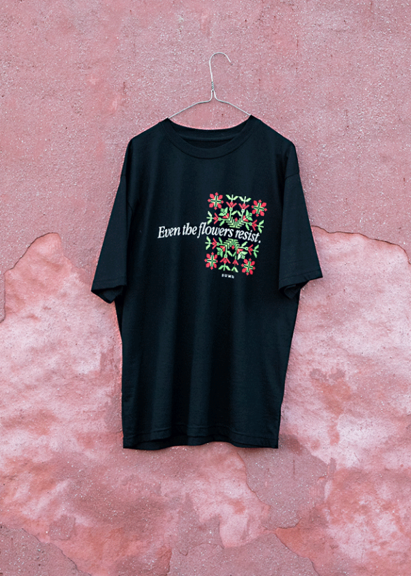 A black T-shirt hangs on a wire hanger against a pink textured wall. The shirt features colorful flowers and the text "Even the flowers resist.