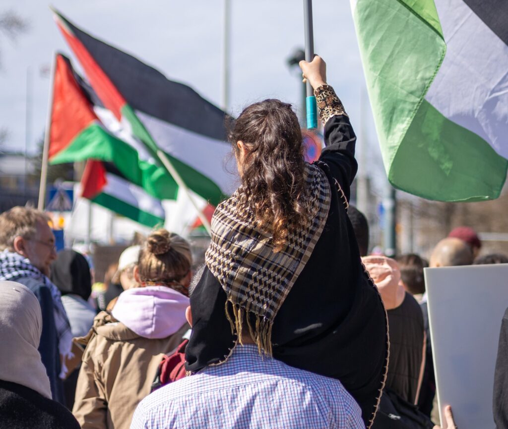 A young woman, draped in a checkered scarf, rides on someone's shoulders during a protest. She holds a flag as part of the large crowd, where people carry colorful flags and signs on a sunny day.