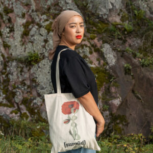 A person wearing a black shirt, blue jeans, and a beige headscarf stands outdoors in front of a rocky background. They hold a white tote bag with a red flower design.