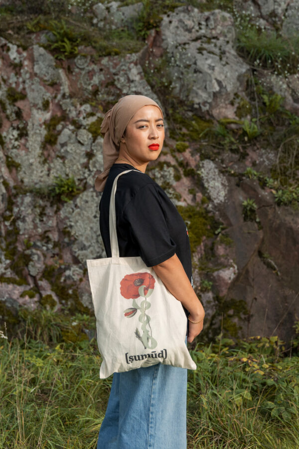 A person wearing a black shirt, blue jeans, and a beige headscarf stands outdoors in front of a rocky background. They hold a white tote bag with a red flower design.