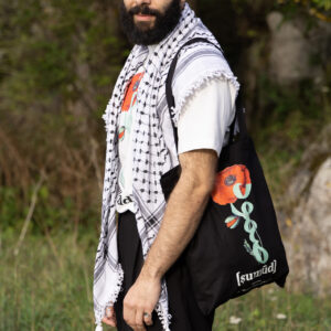 A bearded man stands outdoors wearing a white shirt with a black-and-white patterned scarf. He holds a black tote bag featuring a red flower and Arabic script. He is surrounded by greenery and appears content.