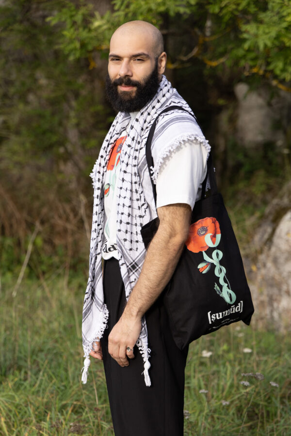 A bearded man stands outdoors wearing a white shirt with a black-and-white patterned scarf. He holds a black tote bag featuring a red flower and Arabic script. He is surrounded by greenery and appears content.