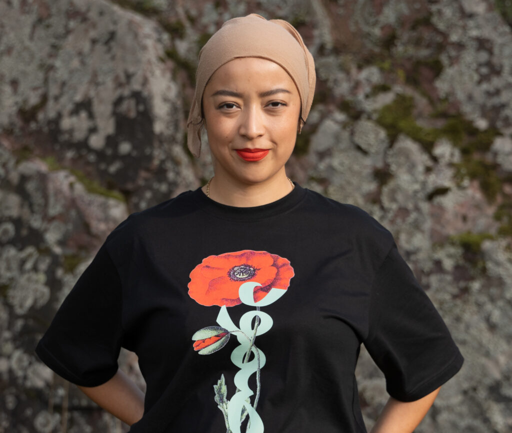 A person wearing a black t-shirt with a large red poppy design stands against a rocky background. They have a beige head wrap and are smiling gently, with hands on their hips.