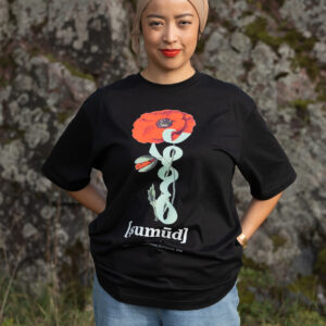 A person wearing a black T-shirt with a red flower design and the word "sumüd" stands in front of a moss-covered rock. They have hands in their pockets, with greenery at their feet.