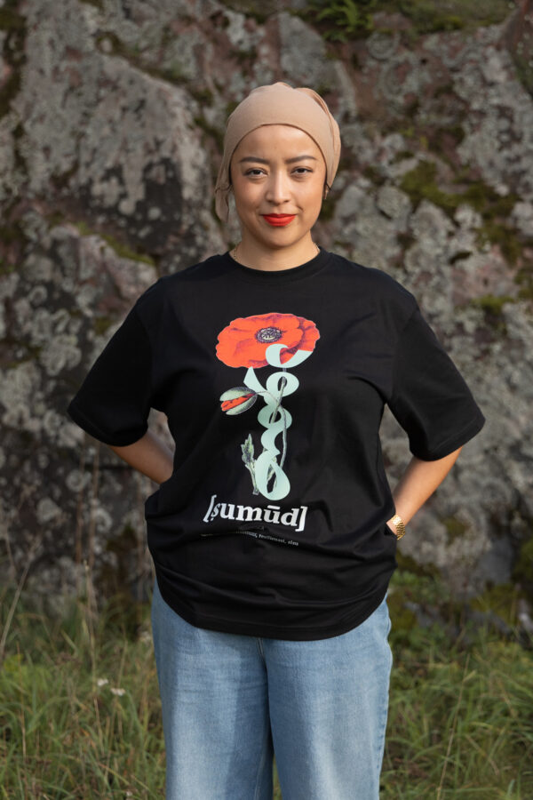 A person wearing a black T-shirt with a red flower design and the word "sumüd" stands in front of a moss-covered rock. They have hands in their pockets, with greenery at their feet.