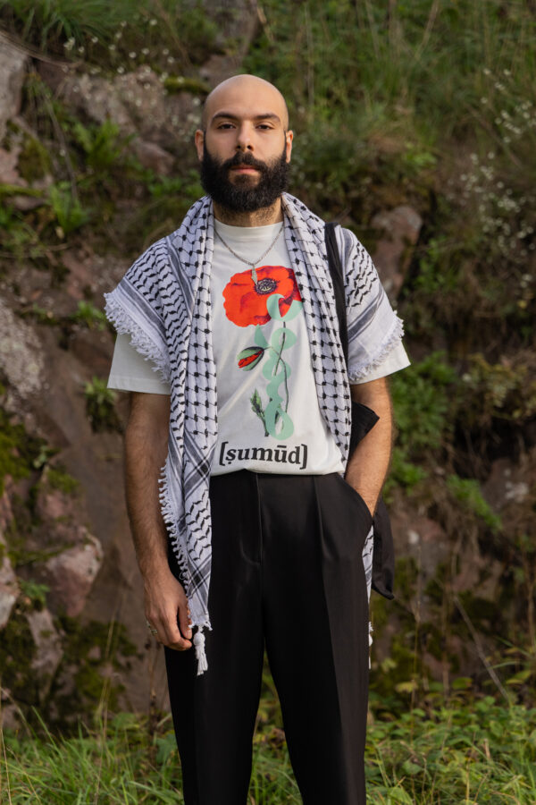 A bearded man stands outdoors in front of rocks and greenery, wearing a patterned scarf and a white T-shirt featuring a red flower and the word "[sumūḍ]." He has his hands in the pockets of his black pants.