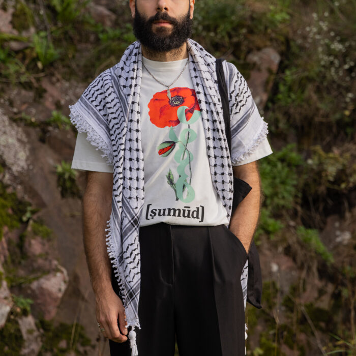 A bearded man stands outdoors in front of rocks and greenery, wearing a patterned scarf and a white T-shirt featuring a red flower and the word "[sumūḍ]." He has his hands in the pockets of his black pants.